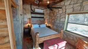 A rustic wooden room with a stone wall, a bed with white linens, a ceiling fan, and a window with blinds. - Cozy, private waterfront stone cabin on the north shore of Lake Travis (Lago Vista)