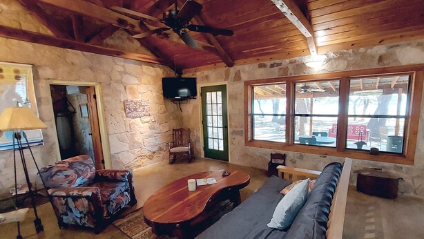 A rustic room with stone walls, wooden ceiling beams, a sofa, a coffee table, a TV, and a window with a view. - Cozy, private waterfront stone cabin on the north shore of Lake Travis (Lago Vista)