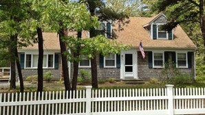 A two-story house with a white picket fence, surrounded by trees. - Experience the charm of Cape Cod, a spacious 4-bedroom retreat in Eastham (Eastham)