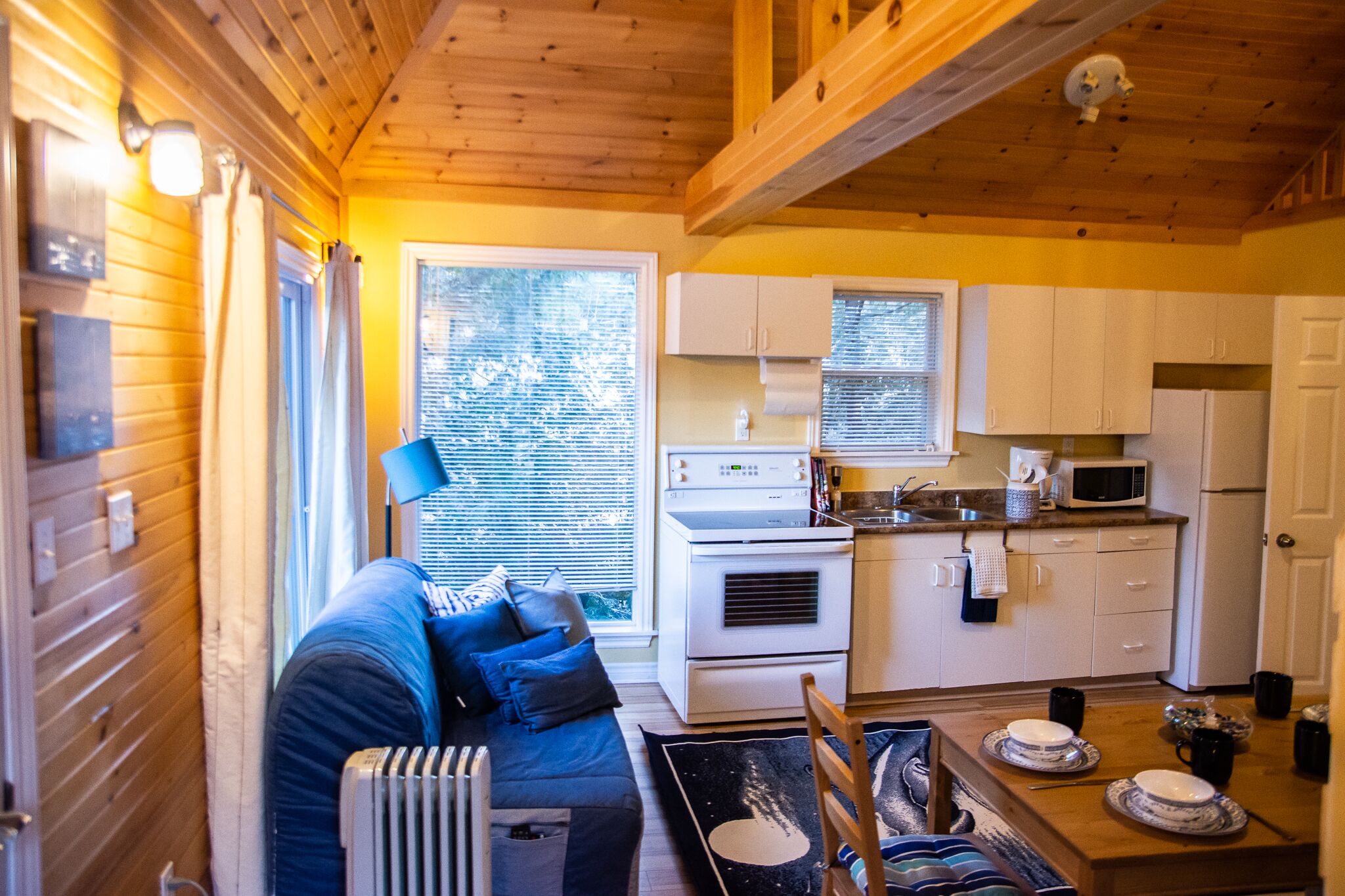 A compact kitchen with white appliances, a dining table with plates, and a blue sofa in a cabin-style room.