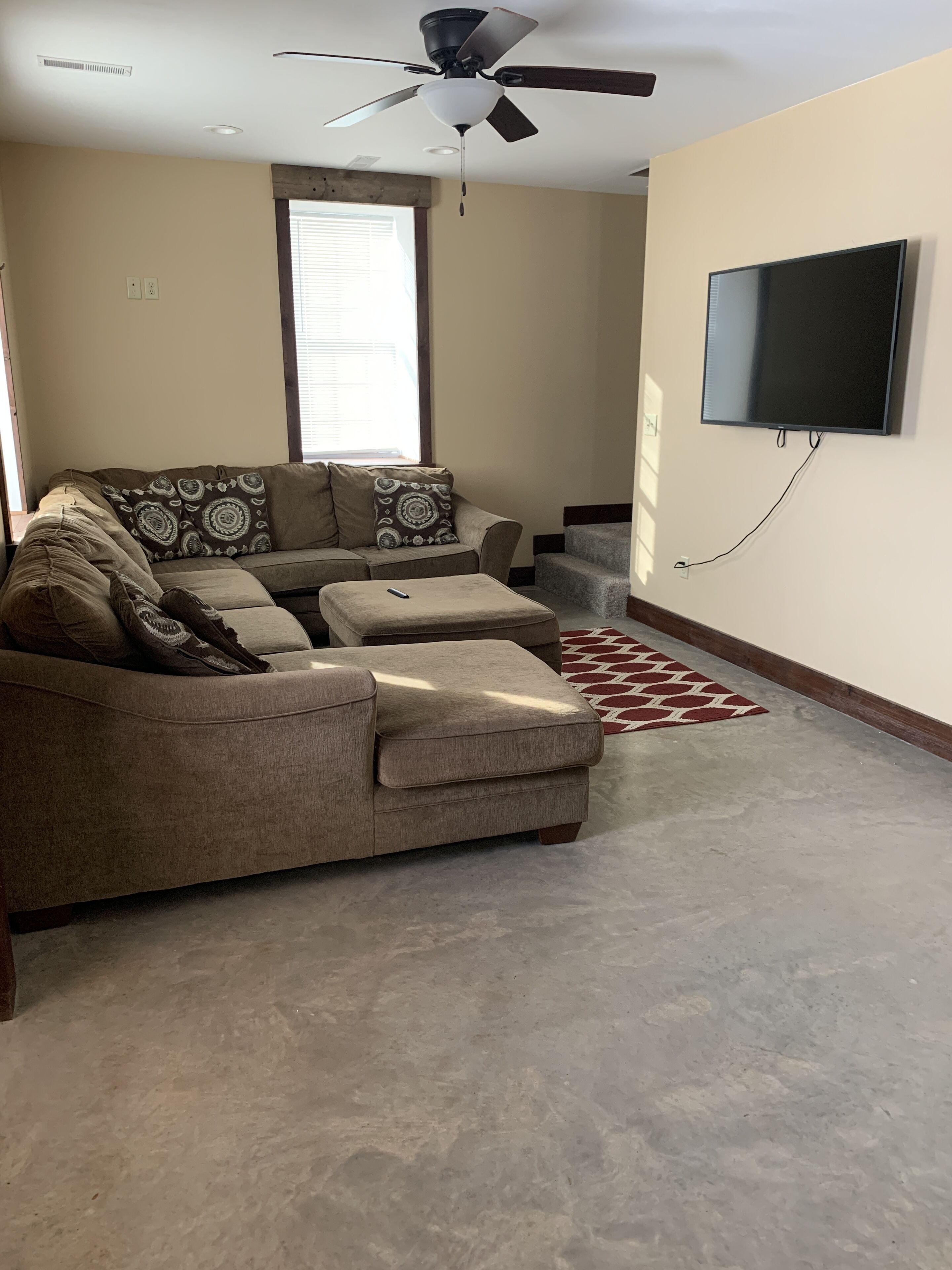 A living room with a sectional sofa, a ceiling fan, a flat-screen TV mounted on the wall, and a concrete floor.