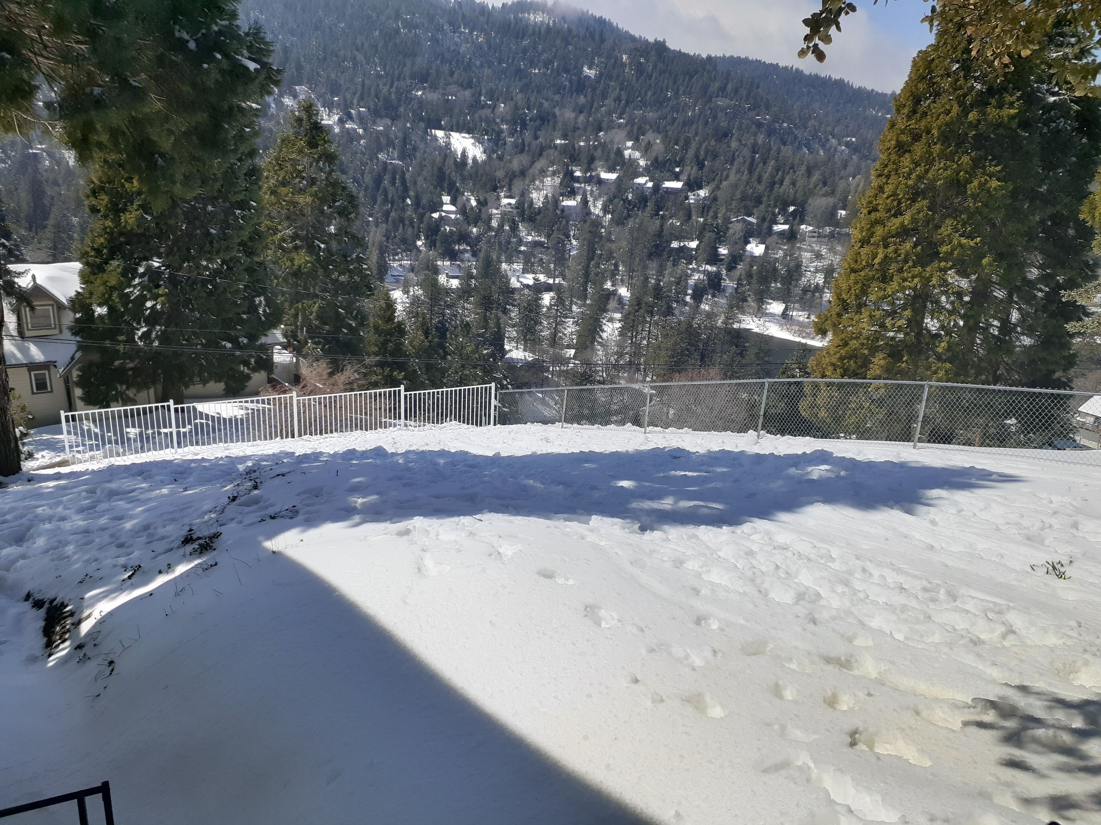 Paesaggio innevato con un'area recintata, alberi e in lontananza case e montagne.