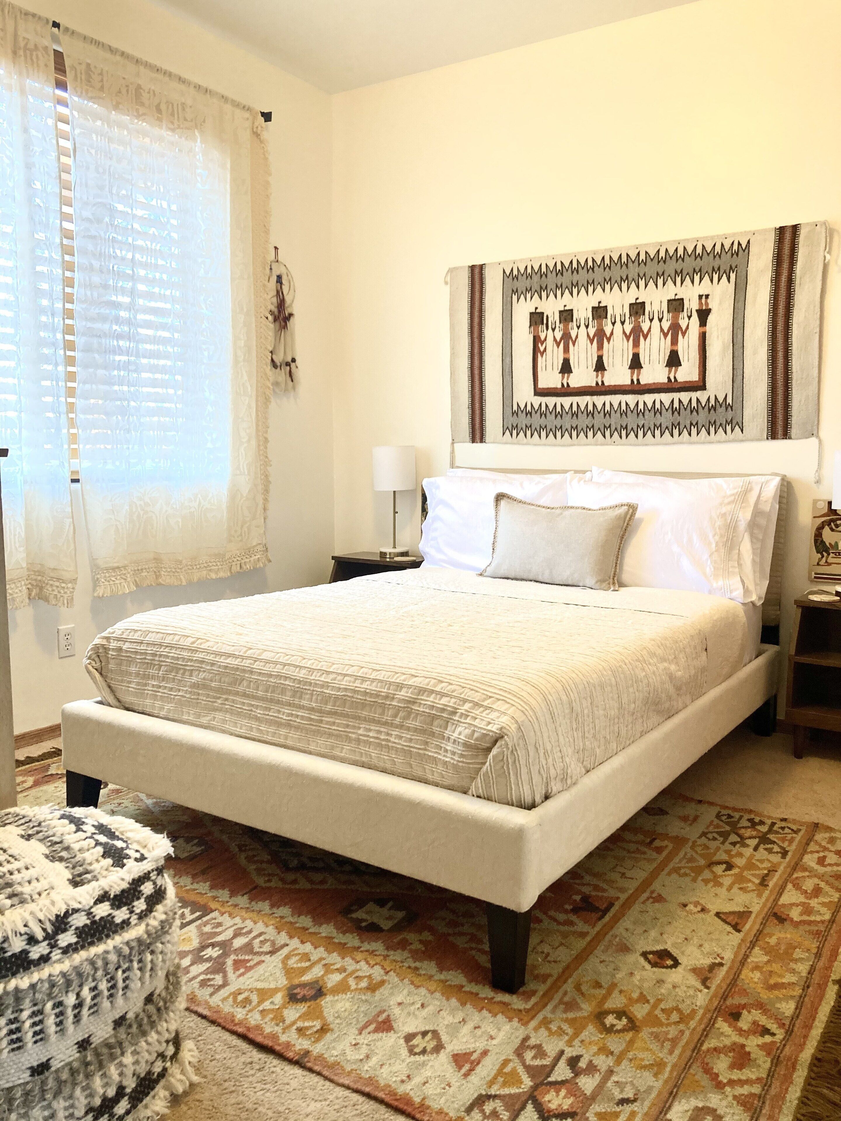 A bedroom with a large bed, a patterned rug, a wall tapestry, and a bedside table with a lamp.