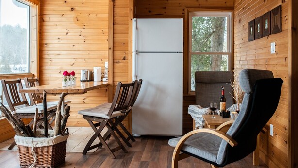 A wooden interior with a dining table, chairs, a refrigerator, and a window with a view of trees. - Lakeview Cottage - Myers Cave Resort (North Frontenac)