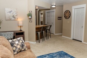 A living room with a sofa, a coffee table, a lamp, and a painting on the wall. There is a dining area with a table and chairs.
