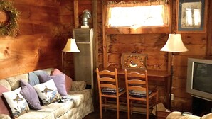 A cozy cabin interior with a wooden ceiling, a sofa with cushions, a chair, a television, and a fireplace with a mantel. - Cottages by the Lake #2   (Bristol)
