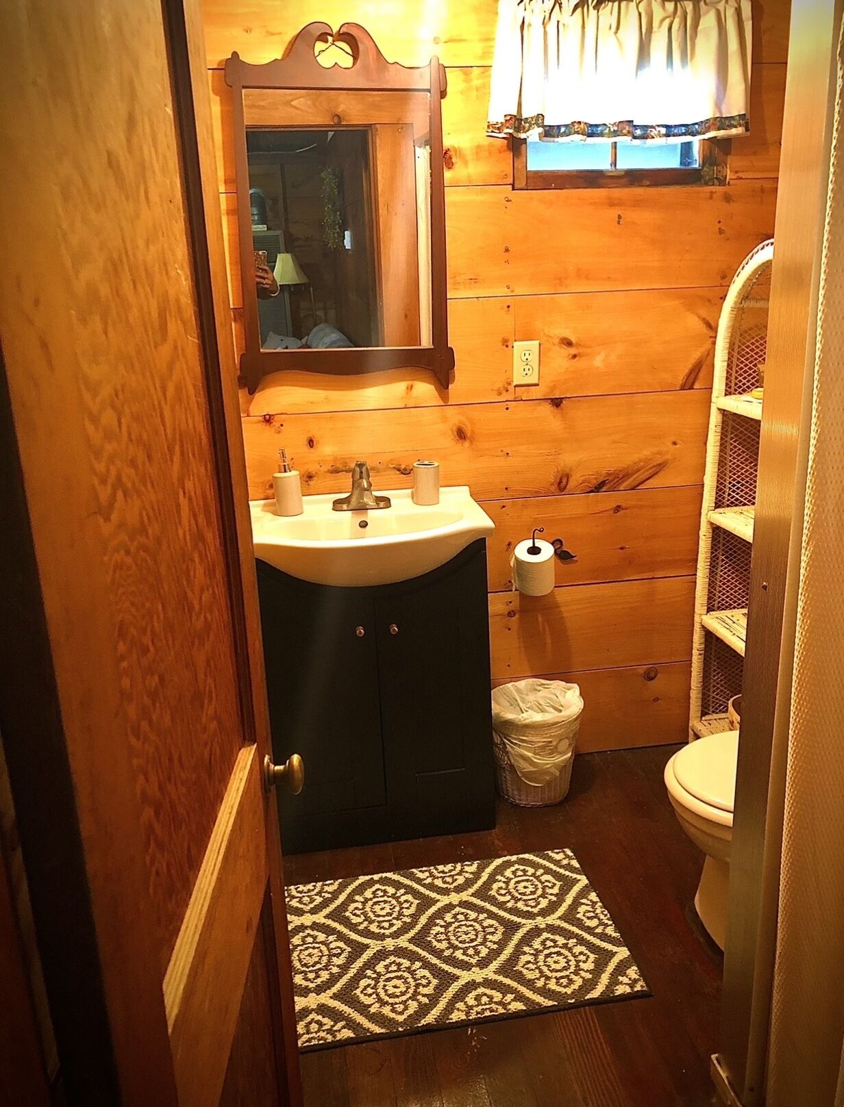 Un baño de madera con lavamanos, espejo, inodoro y un pequeño cubo de basura.