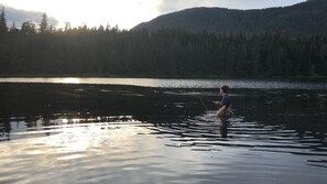 Eine Person angelnd in einem ruhigen See, im Hintergrund ein bewaldeter Berg.