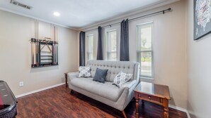 A living room with a grey sofa, two patterned cushions, a wooden side table, and a window with curtains. - Prime Location - Pool Table - BBQ - Walk to AT&T (Arlington)