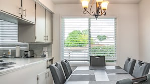 A modern kitchen with a dining area, featuring a large window with blinds, a chandelier, and a table with chairs. - Parkside Estate, Niagara Falls, USA + Free Parking (Niagara Falls)