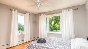 A bedroom with a bed, two windows with curtains, a ceiling fan, and a view of greenery outside. - Parkside Estate, Niagara Falls, USA + Free Parking (Niagara Falls)