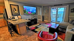 A living room with a TV, a sofa, a red chair, a kitchen area, and a view of the lake through a sliding door.