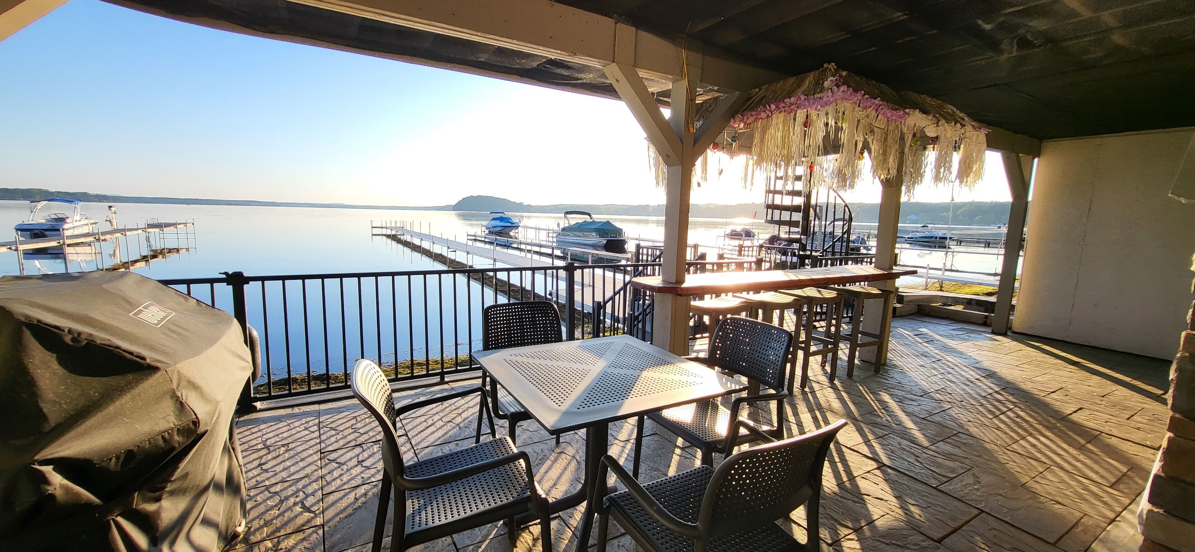 An outdoor patio with tables and chairs, overlooking a calm body of water with boats docked.