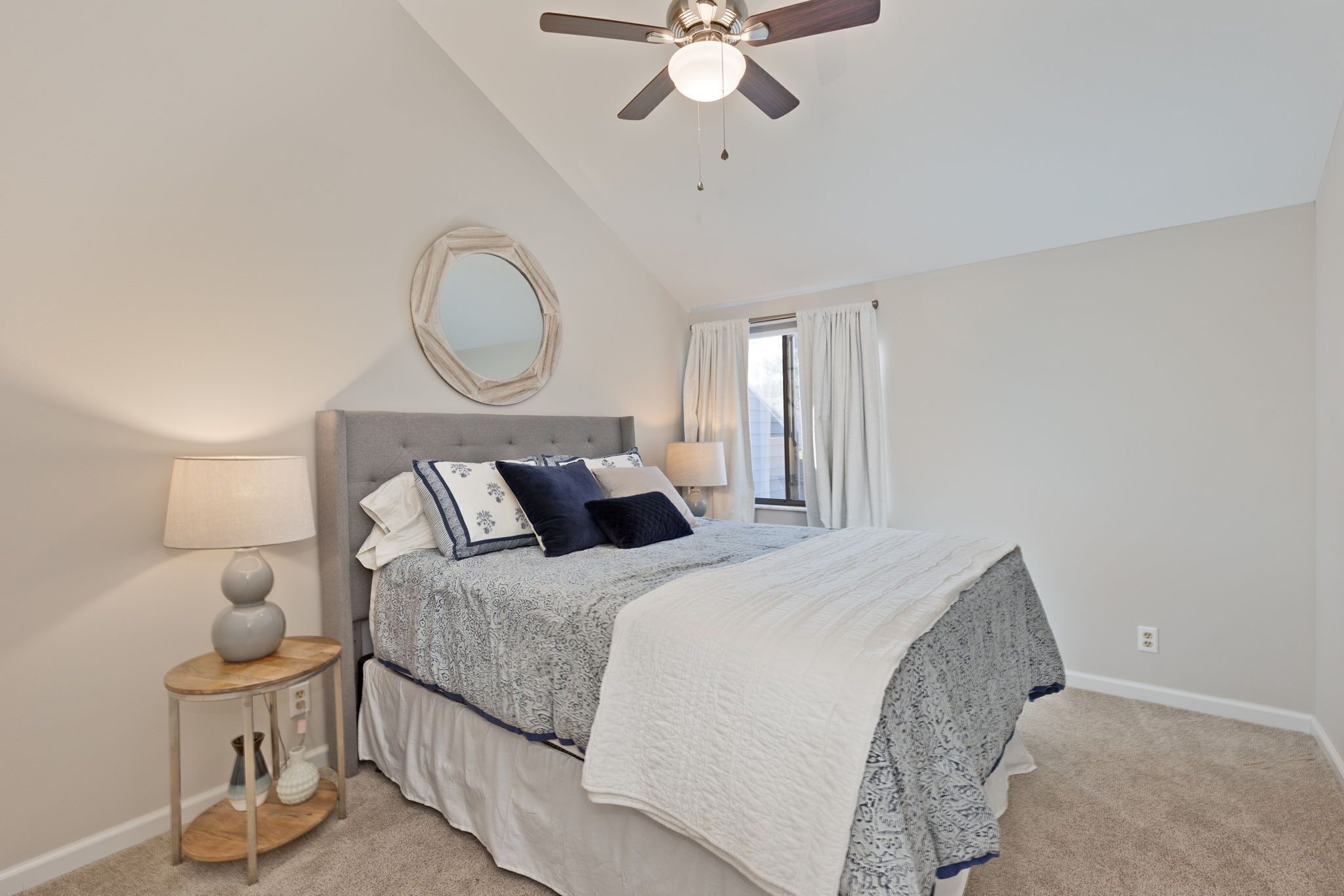 A bedroom with a bed, a round mirror, a ceiling fan, and a window with curtains.