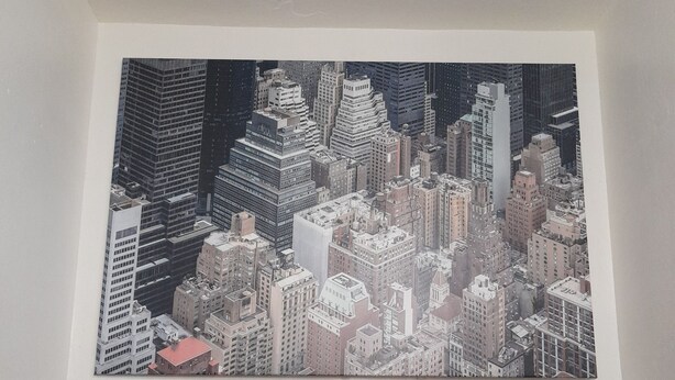 Une peinture représentant un paysage urbain est accrochée au mur, au-dessus d’un canapé marron agrémenté de deux coussins rouges et d’un tableau encadré représentant un paysage.
