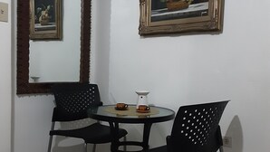 Un petit coin repas avec deux chaises noires, une table ronde, une cafetière et deux tasses. Un miroir et un tableau encadré ornent le mur.