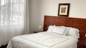 A neatly made bed with a wooden headboard, a framed picture on the wall, and a window with sheer curtains. - Bright 2BR Apartment Steps from El Olivar Park (San Isidro)