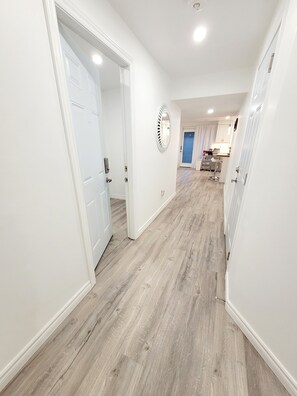 Un couloir aux murs blancs et au plancher en bois, qui mène à une cuisine avec des armoires blanches et une porte bleue.