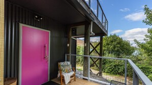 Een houten terras met een roze deur, een stoel met een plaid, en uitzicht op groen en bergen.
