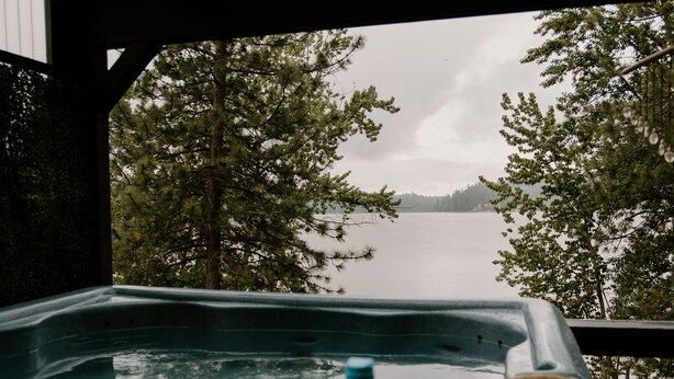 Ein Whirlpool mit Strohhut und Wasserflasche auf einer Holzterrasse mit Blick auf einen See und Bäume.