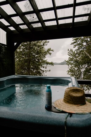 Un bain à remous avec un chapeau de paille et une bouteille d’eau, installé sur une terrasse en bois donnant sur un lac et des arbres.