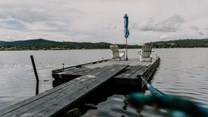 Eine Holzstele ragt in ruhiges Wasser, mit zwei Adirondack-Stühlen und einem blauen Sonnenschirm.