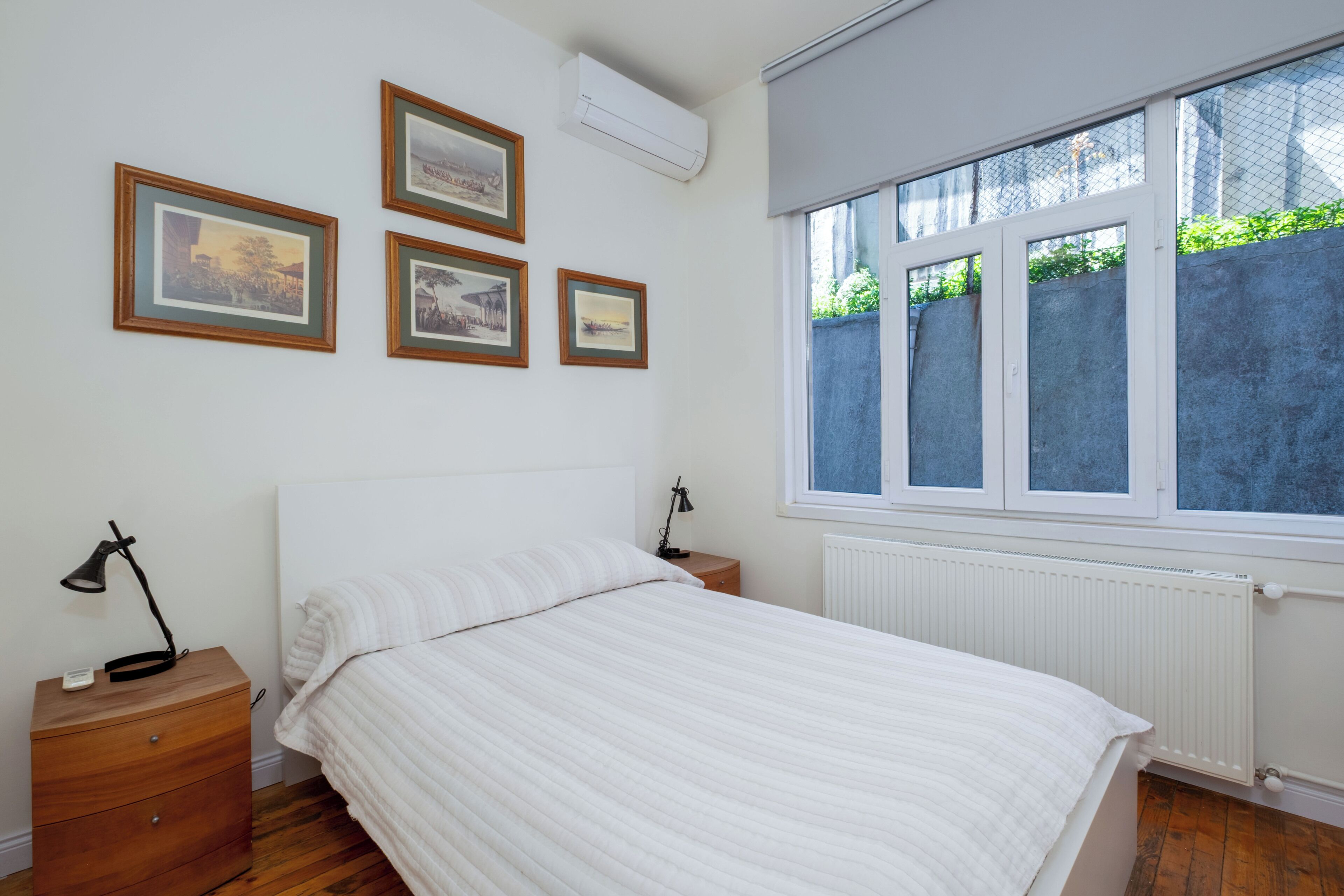 Ein Schlafzimmer mit einem Bett, einem Nachttisch, einer Lampe, einem Fenster und drei gerahmten Bildern an der Wand.