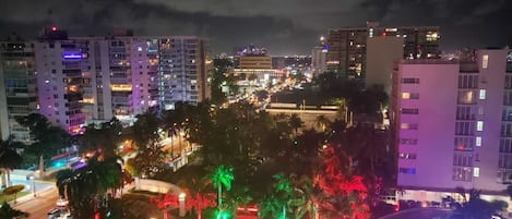 Paesaggio urbano notturno con edifici illuminati, una strada ben illuminata e un’area verde con alberi.