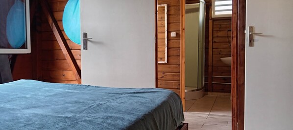 A bedroom with a bed, wooden walls, a door leading to a bathroom, and a wall-mounted light fixture. - Wooden Kaz – Creole accommodation in Le Moule, Guadeloupe (Le Moule)