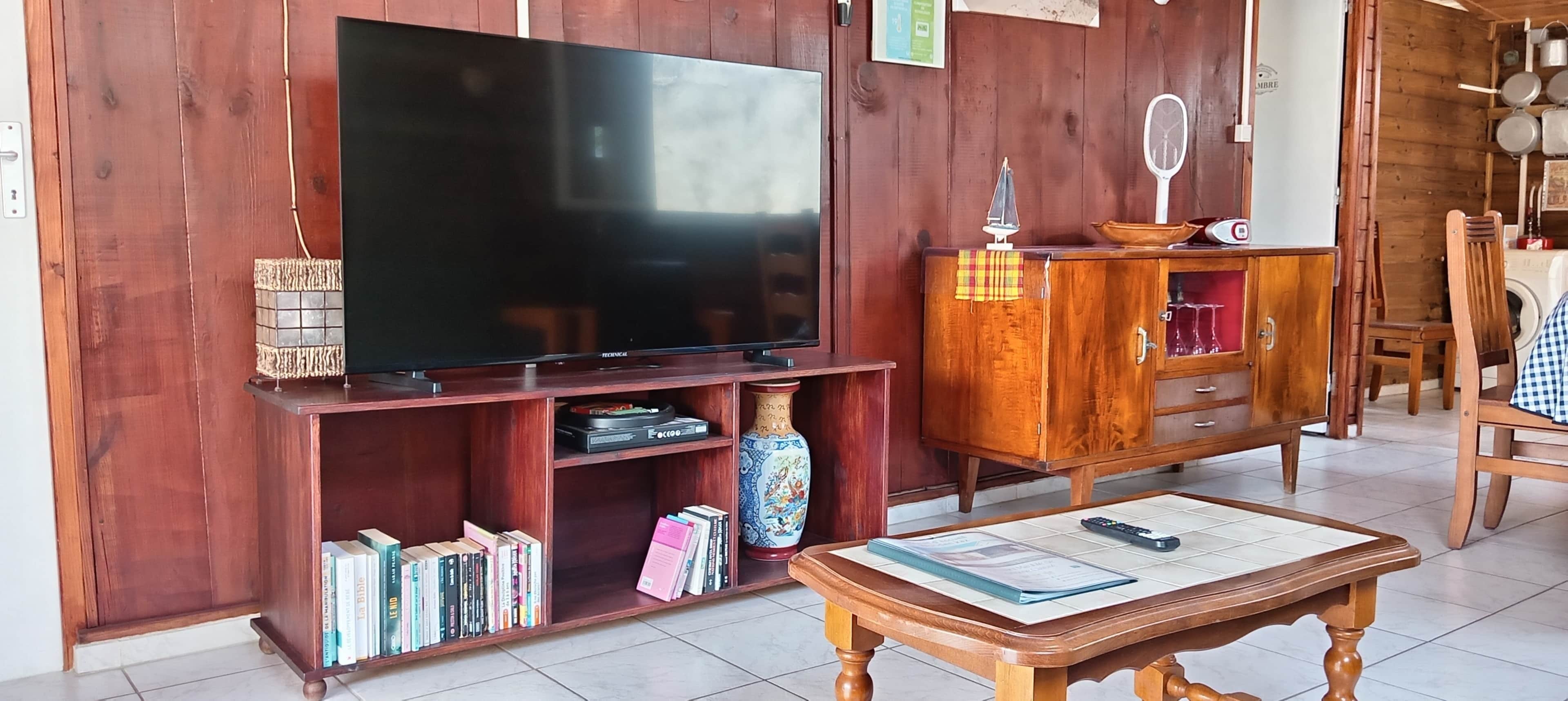 Una sala de estar con una televisión sobre un mueble de madera, una estantería llena de libros, un jarrón decorativo y un aparador con cristalería.
