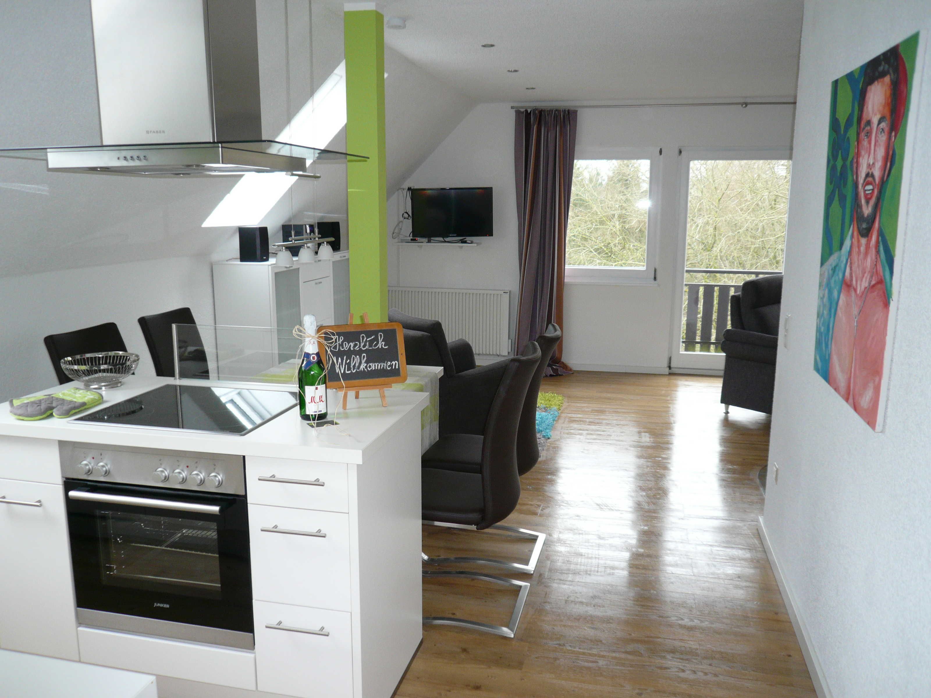 A modern kitchen with a built-in oven, a dining area with a table and chairs, a television mounted on the wall, and a balcony with a view.