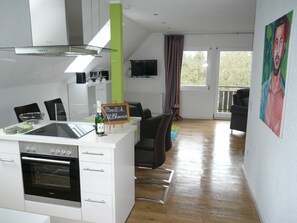 A modern kitchen with a built-in oven, a dining area with a table and chairs, a television mounted on the wall, and a balcony with a view. - Die Wohlfühloase, Deutschland (Frankenau)