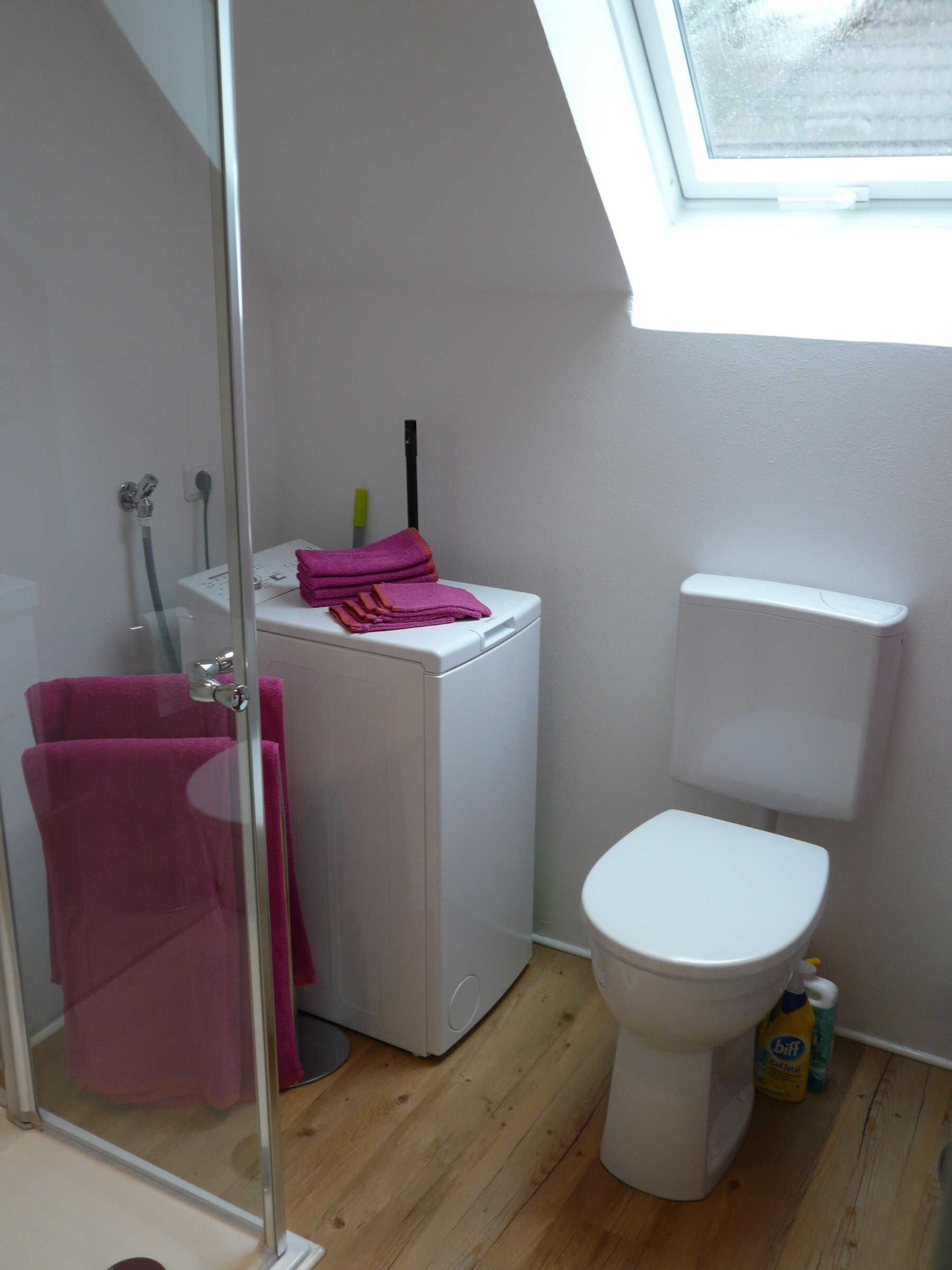 A small bathroom with a glass shower door, a white toilet, a washing machine, and a bottle of cleaning product.