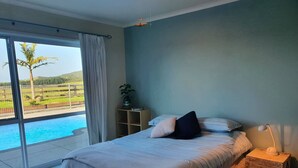 A bedroom with a bed, a nightstand, a lamp, a window with curtains, and a view of a swimming pool and greenery. - Tranquil & private house, outdoor pool, rural setting close to beaches & hikes (Whangarei)