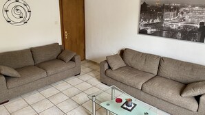 Ein Wohnzimmer mit zwei Sofas, einem Glastisch mit Dekorationsobjekten, einer spiralförmigen Leuchte und einem Stadtbild.