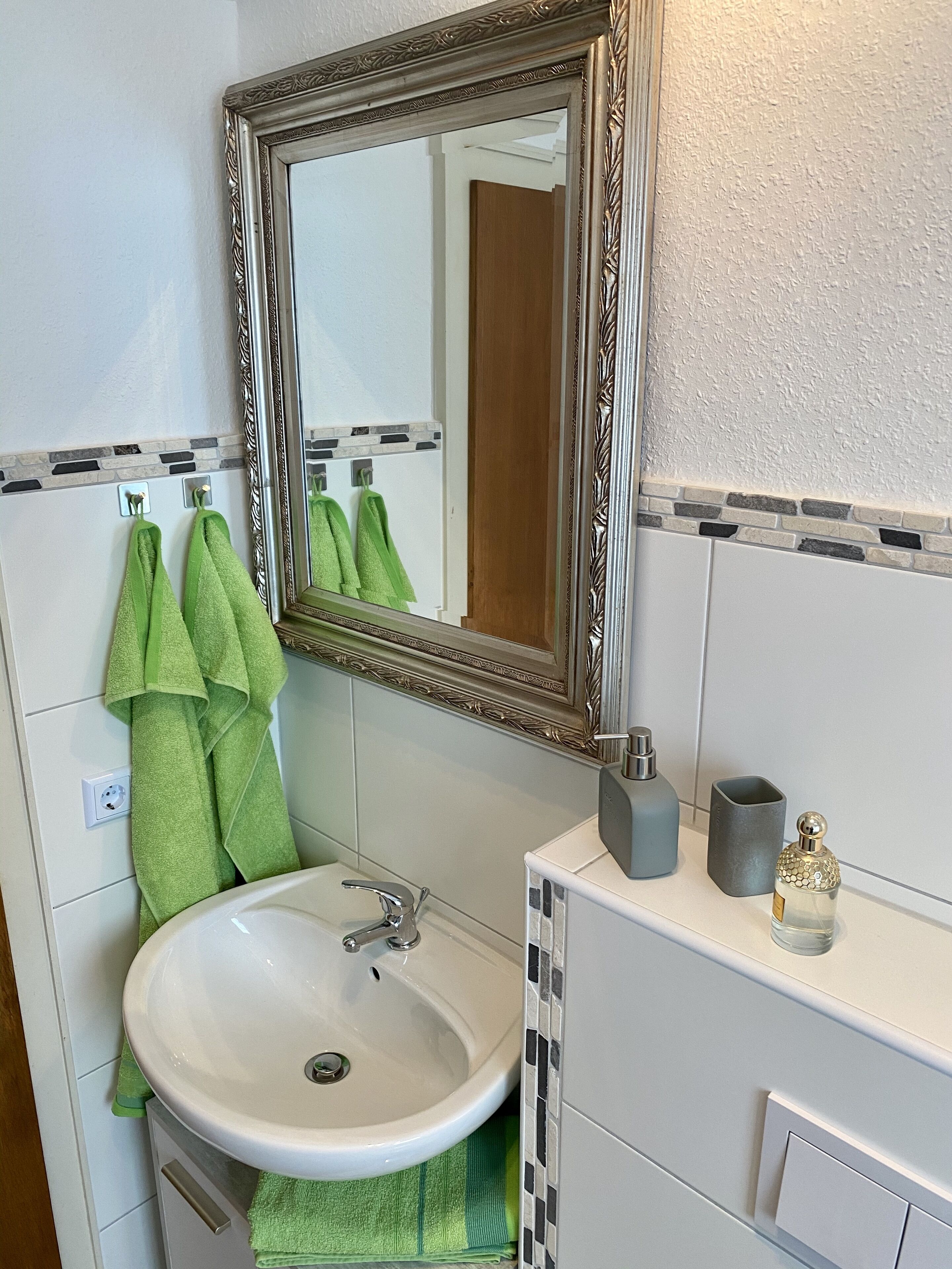 Et badeværelse med en hvid vask, grønne håndklæder, et spejl med en detaljeret ramme og et flisebeklædt backsplash.