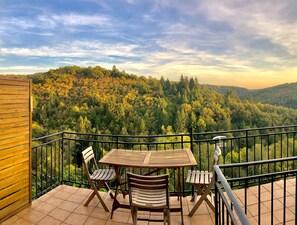 A balcony with a wooden table and two chairs, overlooking a forested hillside.