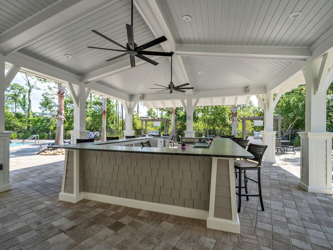 A covered outdoor kitchen area with a central island, ceiling fans, and a view of the pool and greenery.