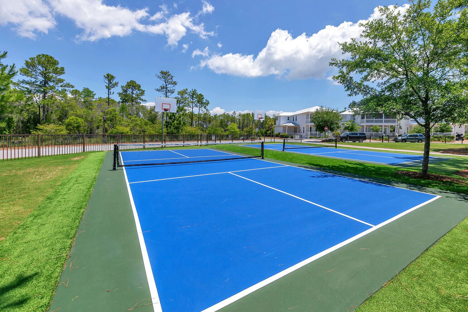A tennis court with a net, surrounded by a fence, trees, and residential buildings in the background.