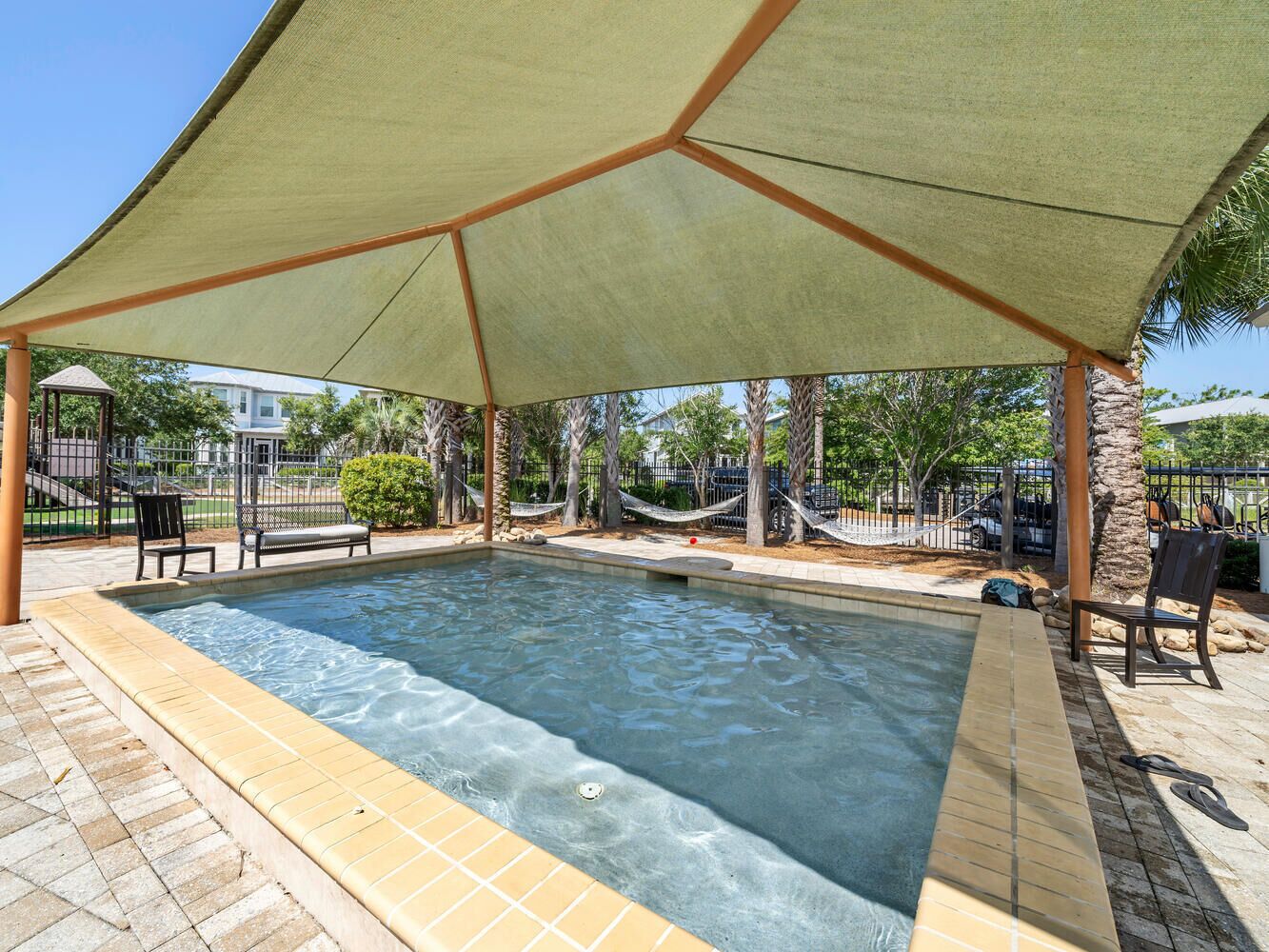 A rectangular swimming pool with a tiled edge, shaded by a large canopy supported by wooden beams.