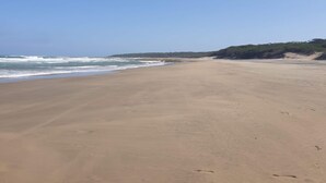 Una amplia playa de arena con olas suaves, cielo despejado y una colina distante cubierta de vegetación.