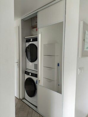 A compact laundry room with a front-loading washer and dryer, a small shelf above the dryer, and a door with a handle. - Modern 4-star 2-bedroom apartment in the center of Aix with parking and terrace (Aix-en-Provence)