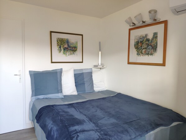 Un dormitorio con una cama, dos cuadros enmarcados, una mesita de noche con una lámpara y una puerta.