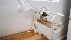 Ein Zimmereck mit einer weißen Schreibtischlampe, einem Korb mit Blumen und einem weißen Metallregal.