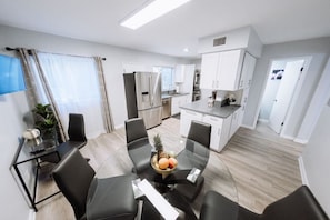 A modern kitchen with stainless steel appliances, a dining area with a glass table and black chairs, and a living area with a TV and curtains.