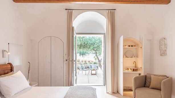 A bedroom with a large bed, a chair, and an open door leading to a balcony with outdoor furniture.