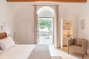 Een slaapkamer met een groot bed, een stoel en een open deur naar een balkon met buitenmeubilair.