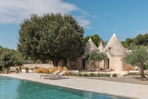 Un bâtiment en pierre avec une piscine, des chaises longues et un grand arbre.