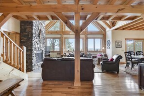 A spacious living area with a stone fireplace, wooden beams, and large windows offering a view of the outdoors. - Walk to the slopes from this beautiful mountain home! Views, Views, Views! (Canaan Valley)