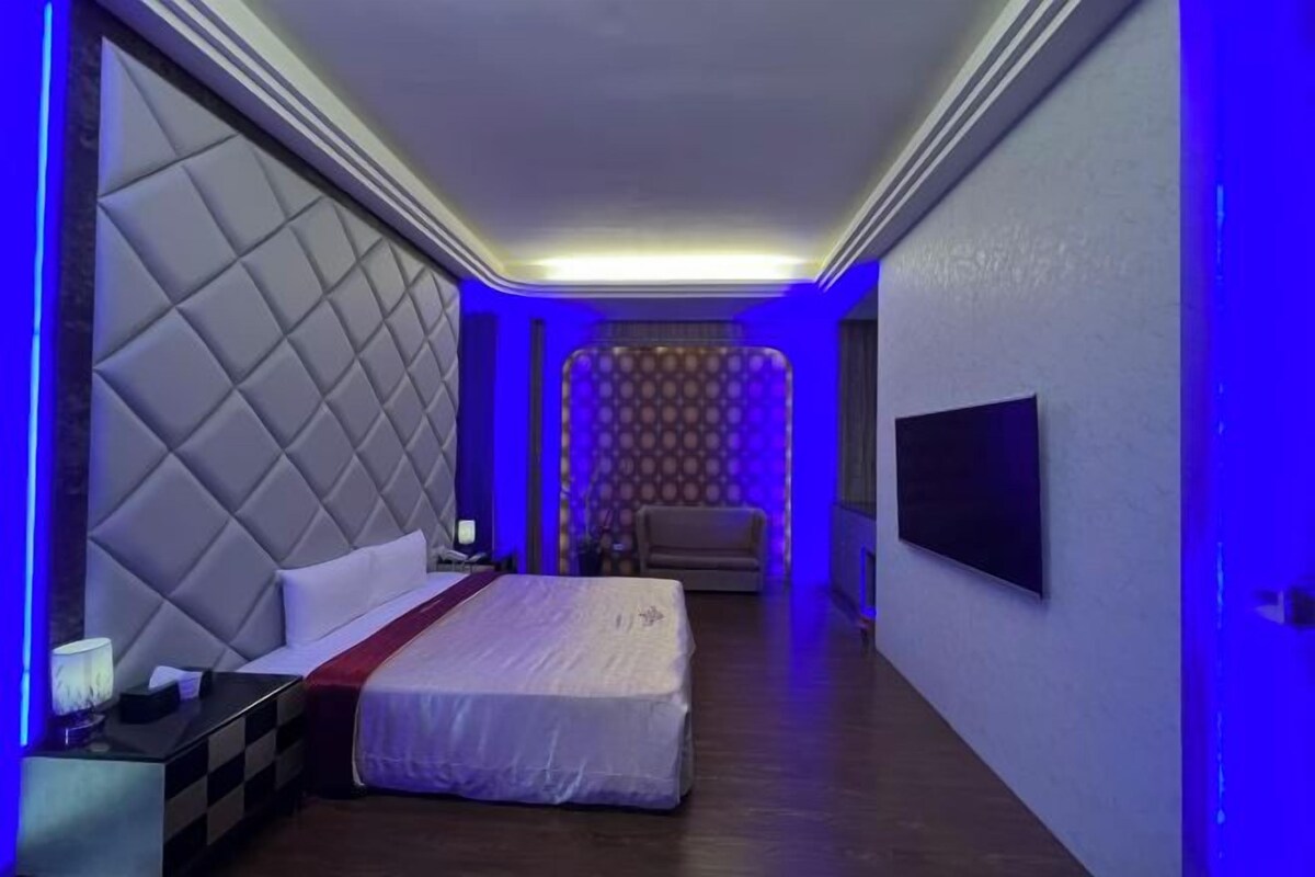 Une chambre d’hôtel avec un grand lit, une table de chevet, un téléviseur fixé au mur et un papier peint à motifs en arrière-plan.